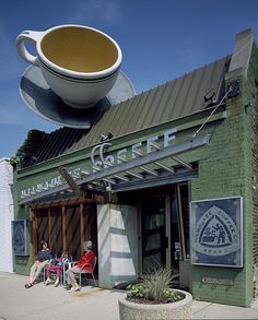 two people sitting on chairs in front of a building with a large coffee cup hanging from it's roof