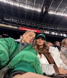 two women sitting next to each other in the stands at a baseball game, one wearing a green hat