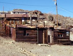 an old wooden building sitting in the middle of a dirt field next to a hill