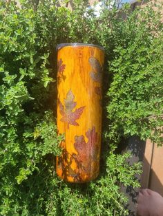 a yellow vase sitting in the middle of some bushes with leaves painted on it's sides