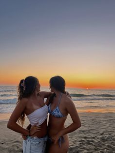 two women in bikinis standing on the beach with their arms around each other as the sun sets