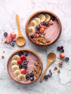 two bowls filled with fruit and granola on top of a marble counter next to spoons