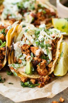 three tacos with meat, cheese and cilantro on parchment paper next to lime wedges