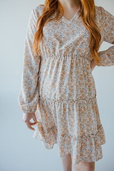 This beautiful neutral midi-length dress has all of the boho vibes! It is going to be your new go-to for date nights, weddings, and work events. This dress features a modest, shorter v-neckline and fluttery blouson long-sleeves. The blousy material flows beautifully down the dress's skirt, with the bottom featuring a tiered effect edged by ruffles. The smocked waist gives the dress an a-line fit that will accentuate your curves beautifully and provide structure to this extra flowy dress. The lin Spring Boho Knee-length Dress With Boho Print, Spring Knee-length Boho Dress, Beige Mid-length Fall Dress, Spring Boho Print Midi Dress, Beige Boho Print Chic Dress, Brown Boho Long Sleeve Dress For Spring, Brown Long Sleeve Boho Dress For Spring, Chic Beige Boho Print Dress, Brown Modest Midi Dress For Spring