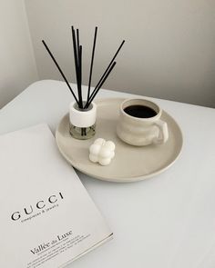 a white plate topped with an open book next to a cup of coffee and reed sticks