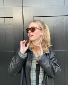 a woman wearing sunglasses standing in front of a wall with her hand on her hip