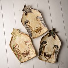 three ceramic ornaments are sitting on a white wooden surface, one has an angel and the other is a nativity scene