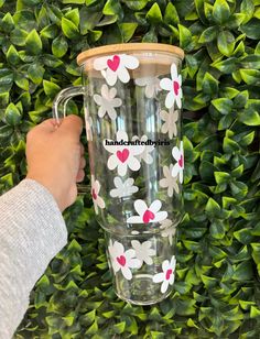 a person holding up a glass cup with flowers on it in front of some bushes