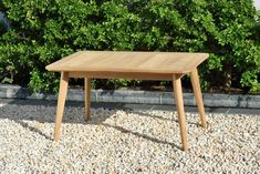 a wooden table sitting on top of a gravel covered ground next to bushes and trees