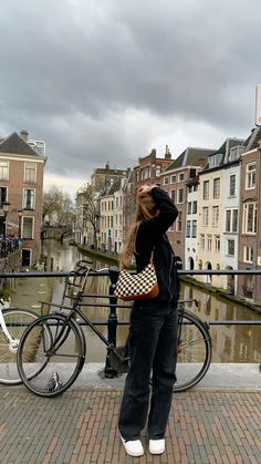 a woman standing next to a bike on a brick walkway near water and buildings with dark clouds in the background