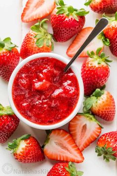 a bowl of strawberry jam surrounded by strawberries on a white surface with a spoon