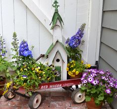 a red wagon filled with potted plants next to a white house and flower pots