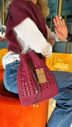 a woman sitting on an orange stool holding a pink crocheted purse with a tag