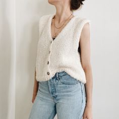 a woman standing in front of a white wall with her eyes closed wearing a sweater and jeans