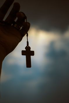 a hand holding a small wooden cross on a chain in front of a cloudy sky