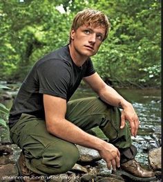 the man is squatting down in front of a river and some rocks with trees behind him