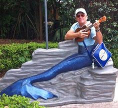 a man sitting on top of a rock next to a blue mermaid holding a guitar