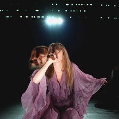 a woman in a purple dress sitting on the ground with her legs spread out and holding a microphone up to her mouth