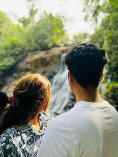 a man and woman standing in front of a waterfall looking at the water from below