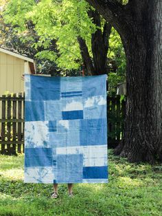 a person holding up a blue and white quilt in front of a tree with green grass