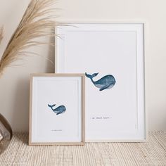 two framed whale prints on a shelf next to a vase with dry grass in it