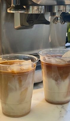 two plastic cups filled with liquid sitting on top of a counter next to an espresso machine