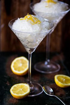 two martini glasses filled with ice and lemon wedges on a black counter top next to silver spoons