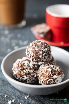 three chocolate coconut balls in a white bowl on a table next to a cup of coffee