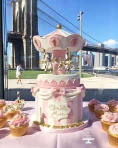 a pink and white carousel cake surrounded by cupcakes on a table in front of a window