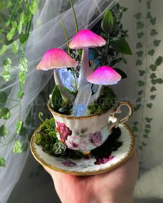 a hand holding a tea cup with pink flowers in it and three mushrooms growing out of the top