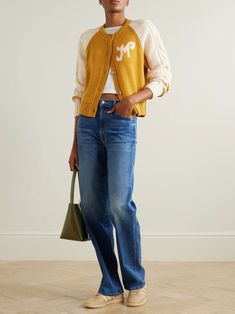 a woman standing in front of a white wall wearing jeans and a yellow cardigan