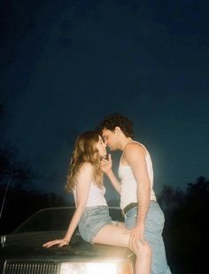 a man and woman sitting on the hood of a car in front of a dark sky