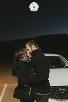 a man and woman kissing in front of a white car at night with the moon above them