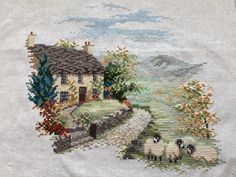 a cross stitched picture of sheep grazing in front of a house with trees and flowers