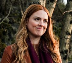 a woman with long red hair wearing a brown jacket and purple scarf standing in front of trees