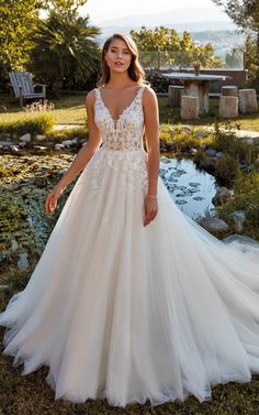 a woman in a white wedding dress standing on the grass near some water and trees