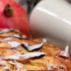 an apple pie with powdered sugar on top and two red apples in the background