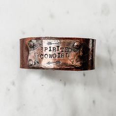 Spirited Cowgirl bracelet is a handmade beauty any free spirited boho cowgirl will boldly and proudly wear. This showpiece has a gorgeous medallion featuring stamped cowgirl symbols including arrows, horse, feather and steer head as well as hand stamped lettering. Handmade in Idaho by Weathered Soul exclusively for Bourbon Cowgirl. Limited Edition, high quality jewelry that is as unique and authentic as you! Due to their handcrafted nature, slight variations will occur. The feel and design of th Rustic Concho Jewelry For Rodeo, Adjustable Stamped Bracelets, Bohemian Hand Tooled Jewelry For Rodeo, Rustic Distressed Brown Bracelet For Gift, Handmade Distressed Brown Adjustable Bracelet, Rustic Hand Tooled Adjustable Bracelets, Hand Tooled Brown Jewelry For Rodeo, Brown Hand Tooled Jewelry For Rodeo, Rustic Adjustable Jewelry For Rodeo