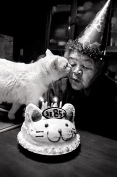 an old woman blowing out the candles on her birthday cake with a cat standing next to her