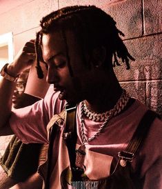 a man with dreadlocks and a pink t - shirt is leaning against a brick wall