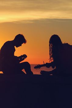 two people sitting on the ground playing guitar at sunset with one person holding a cell phone