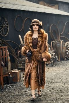 a woman in a fur coat and hat walks down a dirt road near an old building