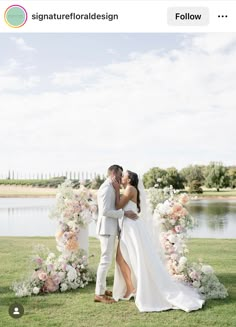 a bride and groom kissing in front of a floral arch on the grass by a lake