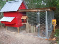 a red chicken coop in the woods behind a chain link fence with an open door