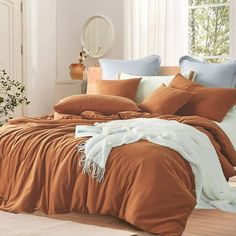 a bed with orange sheets and pillows in a room next to a window, near a vase filled with flowers