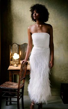 a woman standing in front of a chair wearing a white dress with feathers on it