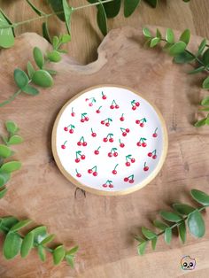 a white plate with cherries painted on it sitting next to green leaves and plants