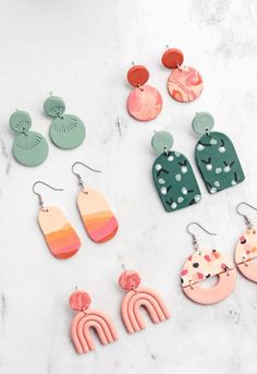 six pairs of colorful earrings on a marble surface with white and green accents, including one in the shape of a cactus