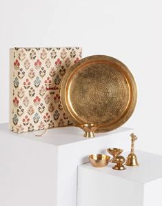 a gold plate and two brass cups on top of a white shelf next to a book