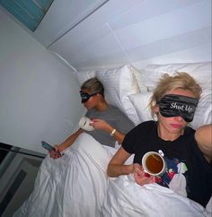 two women laying in bed with blindfolds over their eyes, one holding a cup of coffee